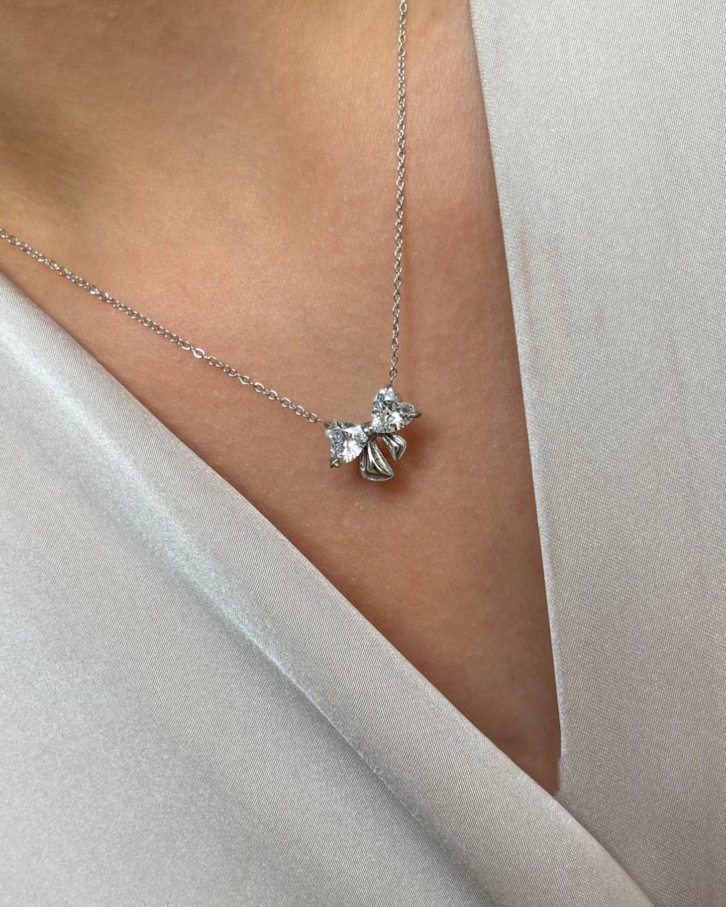 Edelstahl Halskette mit Schleifen Design in silber mit Herzförmigen Zirkonia Steinen. Wasserfest und hypoallergen., getragen mit einer cremefarbenen Bluse.
