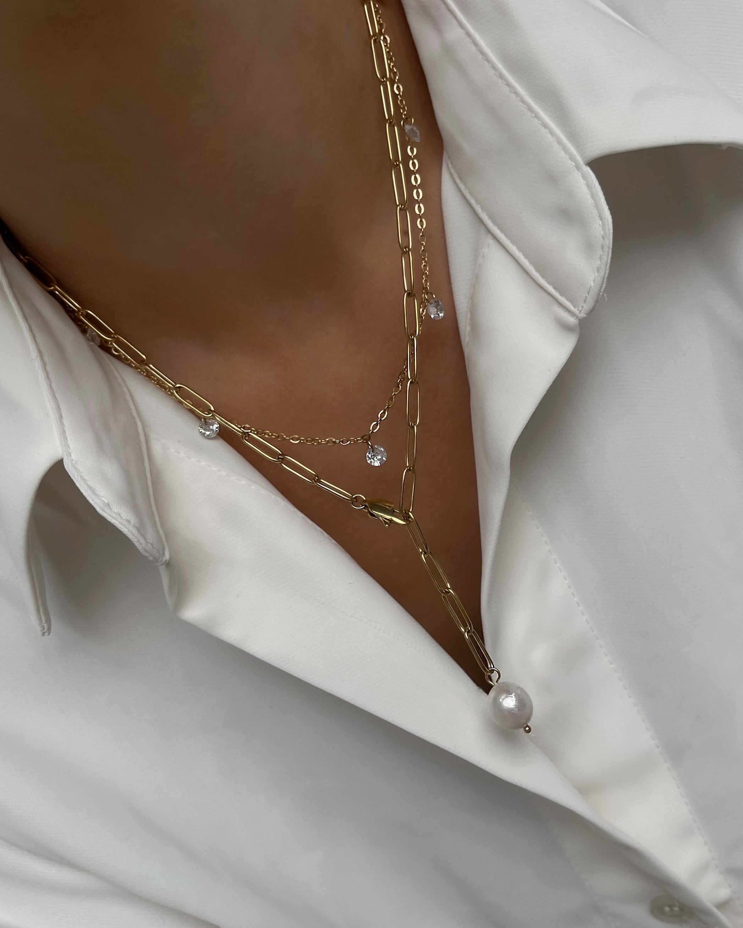 Zwei goldene Halsketten mit sechs kleinen runden Zirkonia Steinen und Perle, getragen mit einer weißen Bluse.