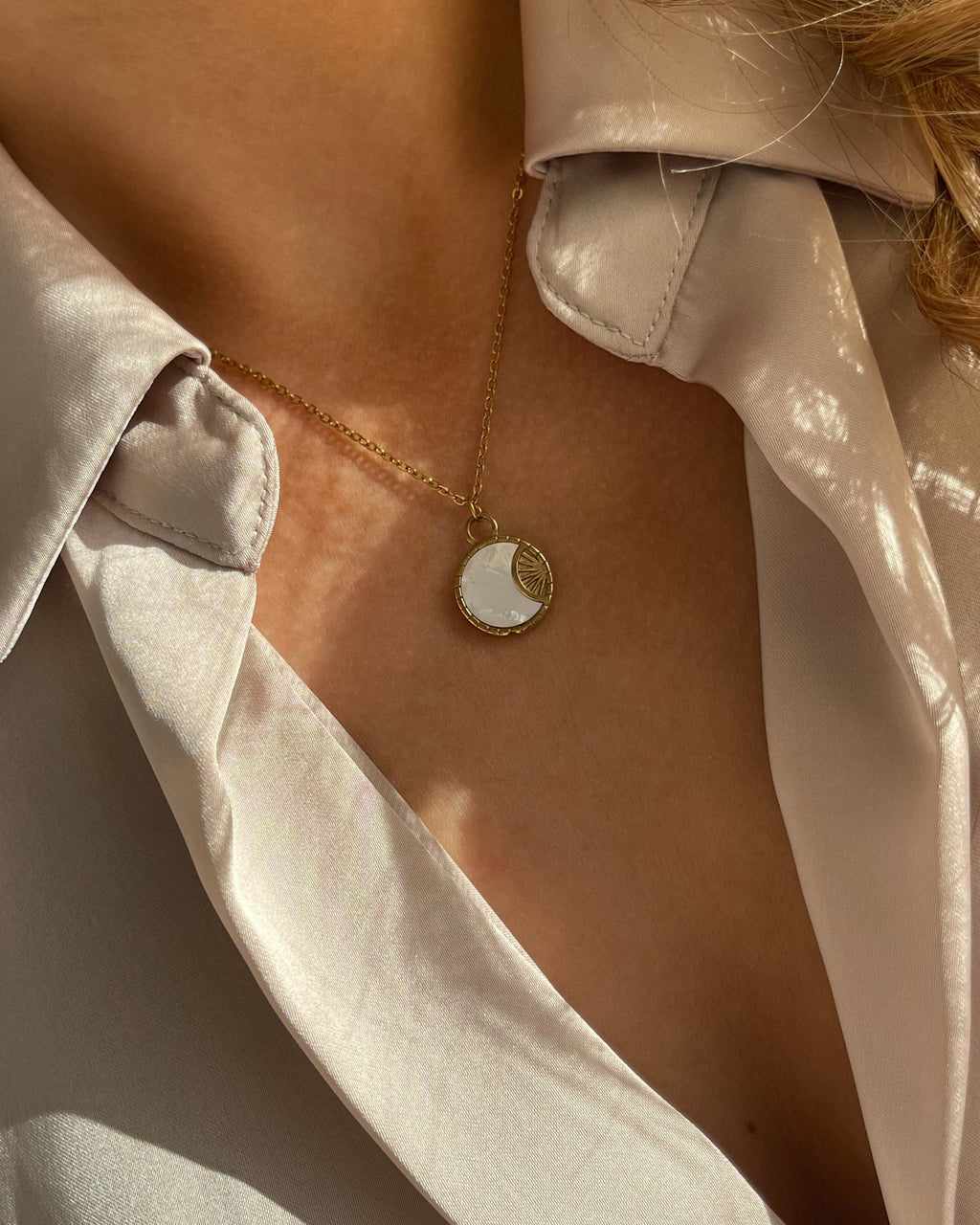 Goldene Halskette mit rundem schimmernden Plättchen, getragen mit beigefarbener Bluse.