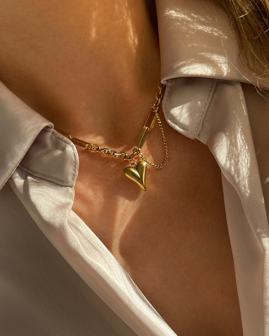 Vergoldete Edelstahlkette mit herzförmigem Anhänger, getragen über einer cremefarbenen Bluse. Wasserfest, hypoallergen und elegant im Design