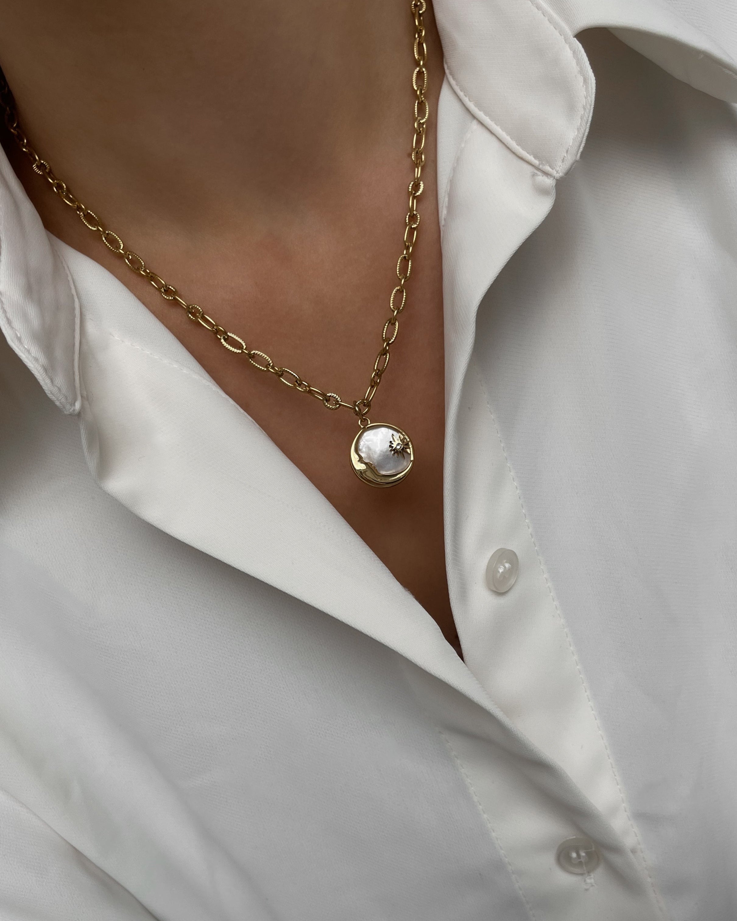Grobe Halskette mit einem Anhänger aus schimmernder Muschelperle und Details in Mond und Sternform, getragen mit einer weißer Bluse.