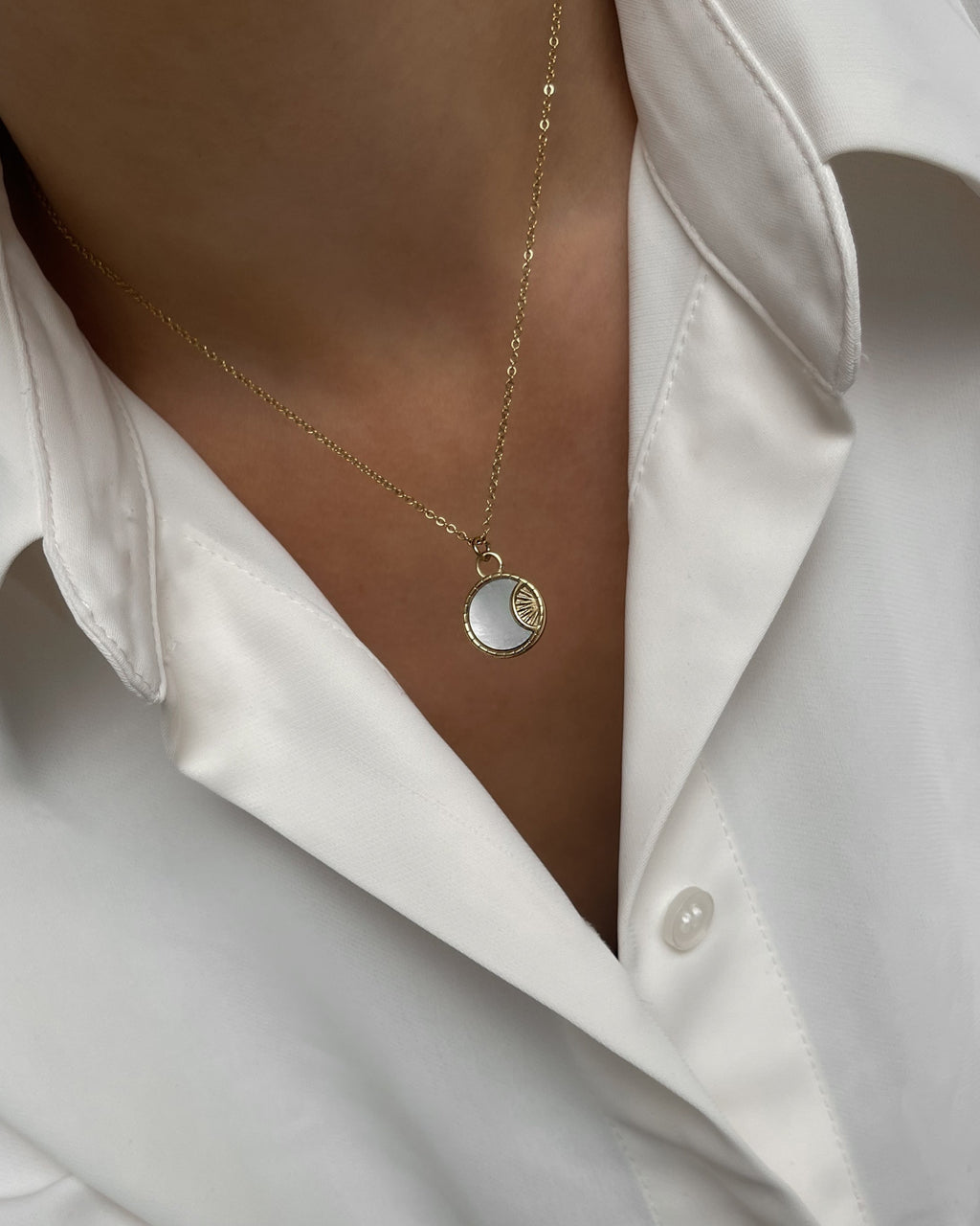 Goldene Halskette mit rundem schimmernden Plättchen, getragen mit weißer Bluse.