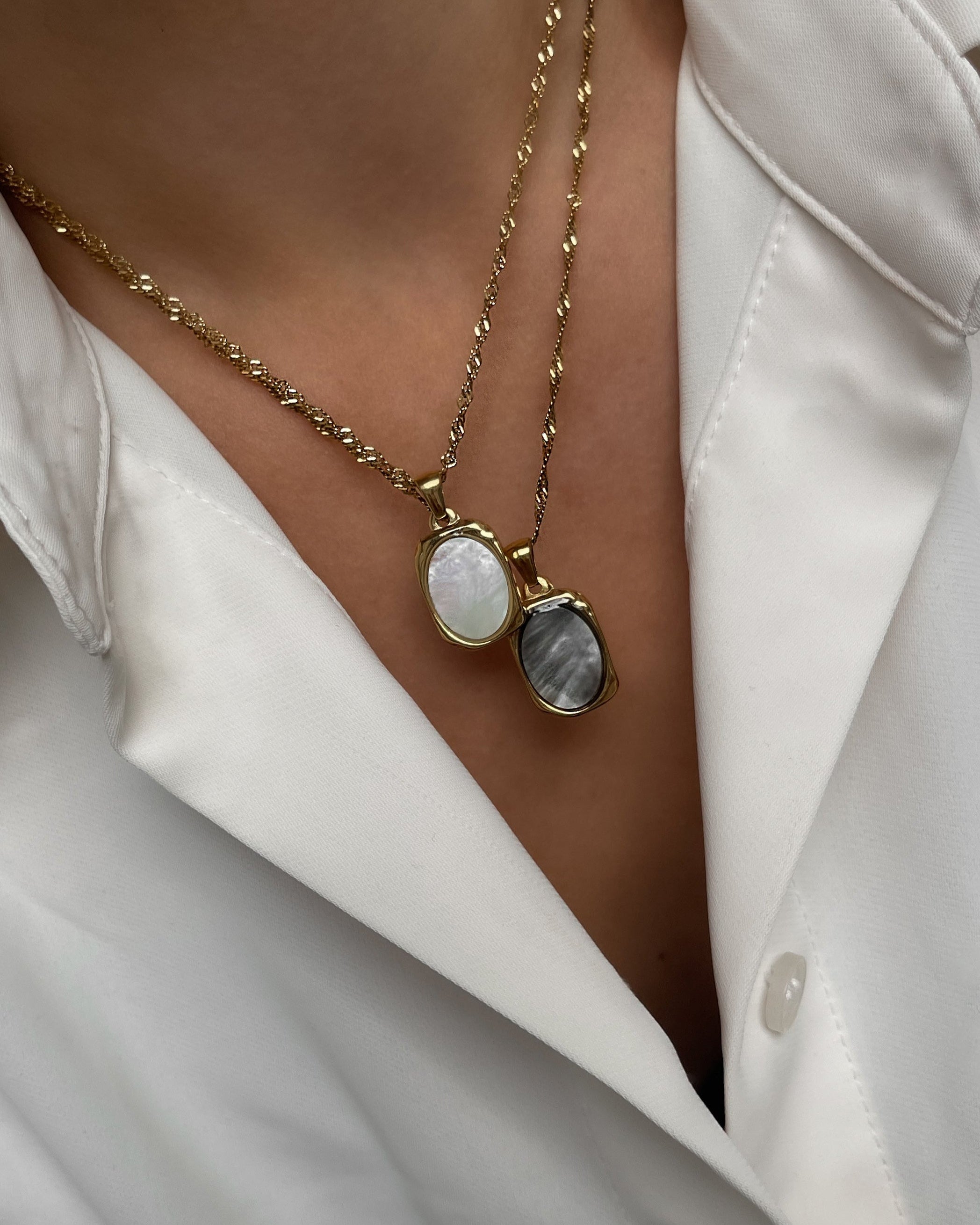 Goldene Halsketten mit ovalem Anhänger aus weißer und grauer Muschelperle, getragen mit weißer Bluse.