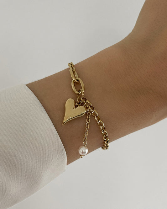 Goldenes Armband mit liebevollem Herz Anhänger und kleinen Perle, getragen am Handgelenk.
