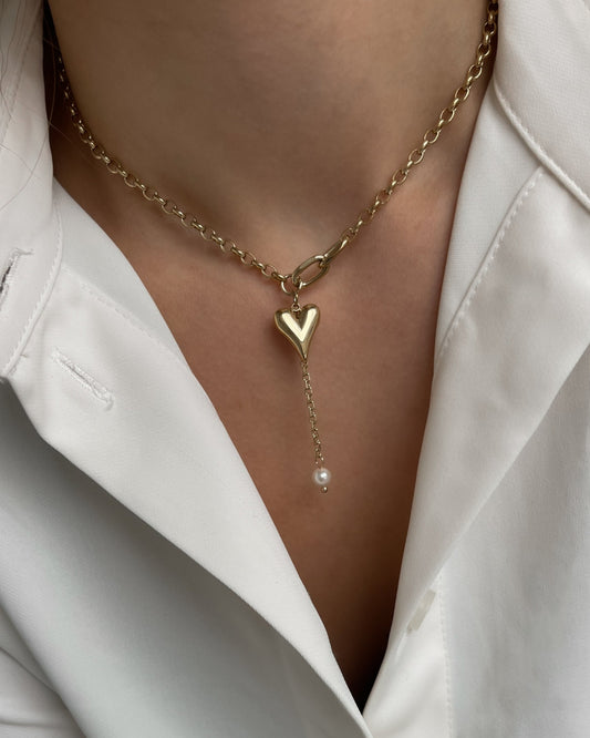 Verspielte goldene Kette mit Herz und Perle, getragen mit einer weißen Bluse.