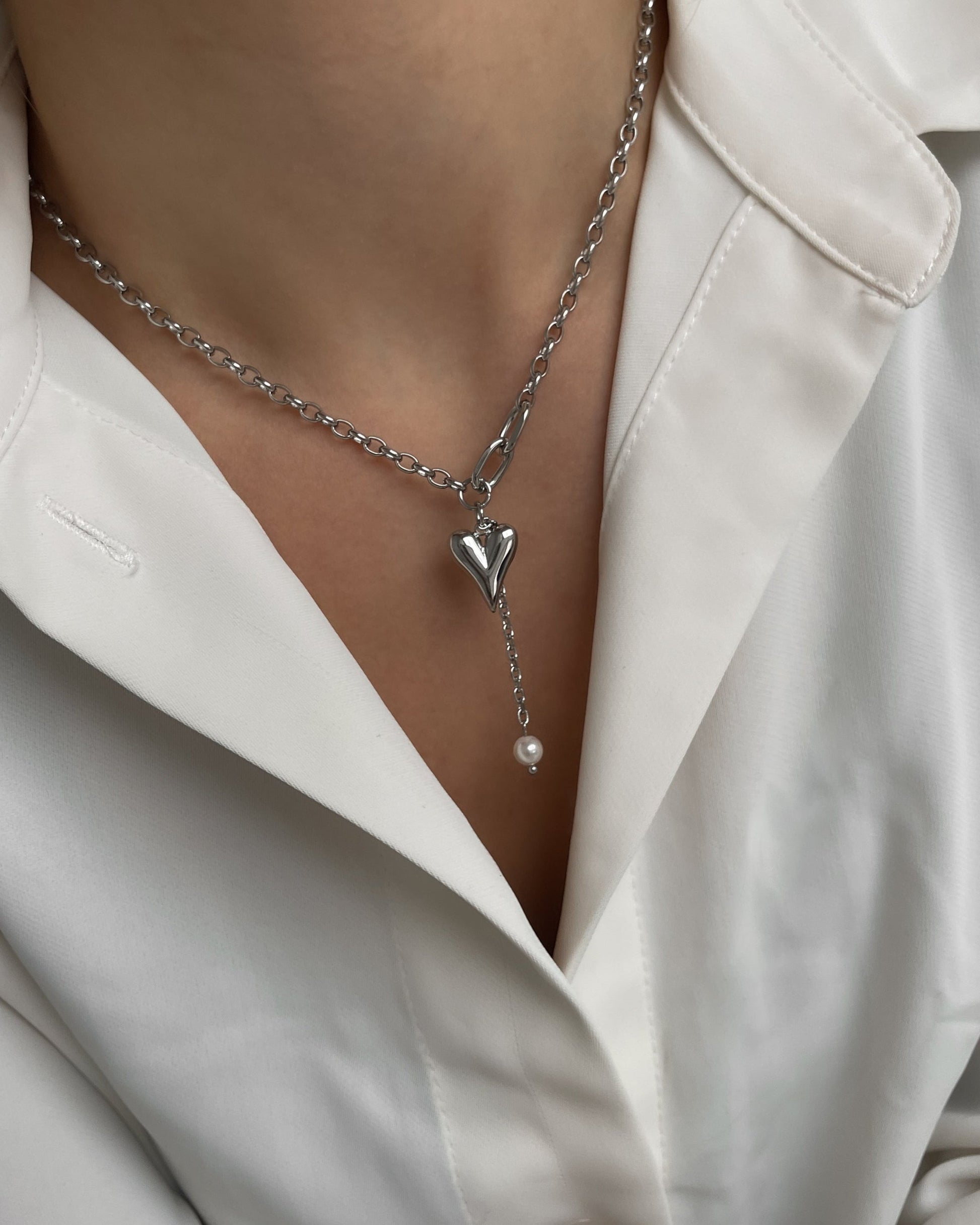 Verspielte silberne Kette mit Herz und Perle, getragen mit einer weißen Bluse.