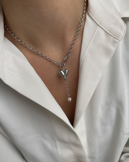 Verspielte silberne Kette mit Herz und Perle, getragen mit einer weißen Bluse.
