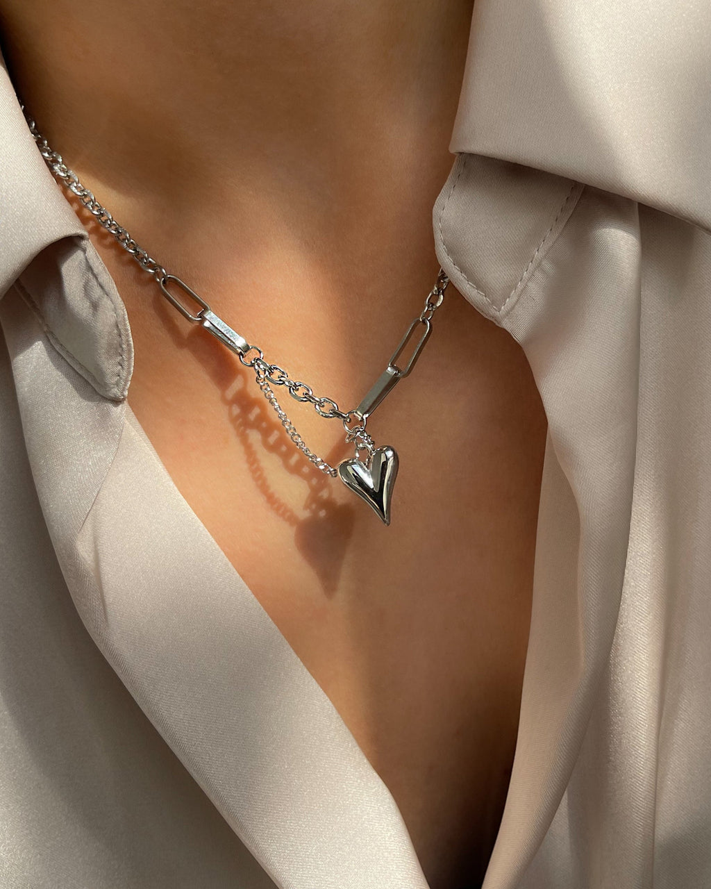 Edelstahlkette mit herzförmigem Anhänger, getragen über einer cremefarbenen Bluse. Wasserfest, hypoallergen und elegant im Design.